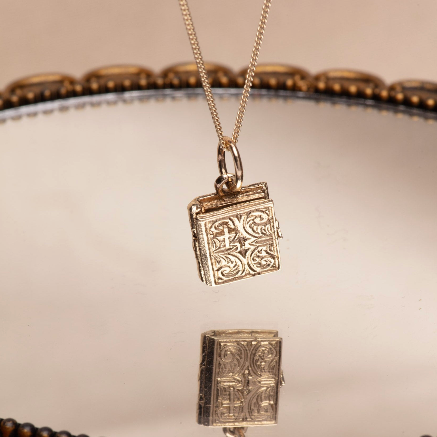 Gold book-shaped pendant on a chain against a neutral reflective background
