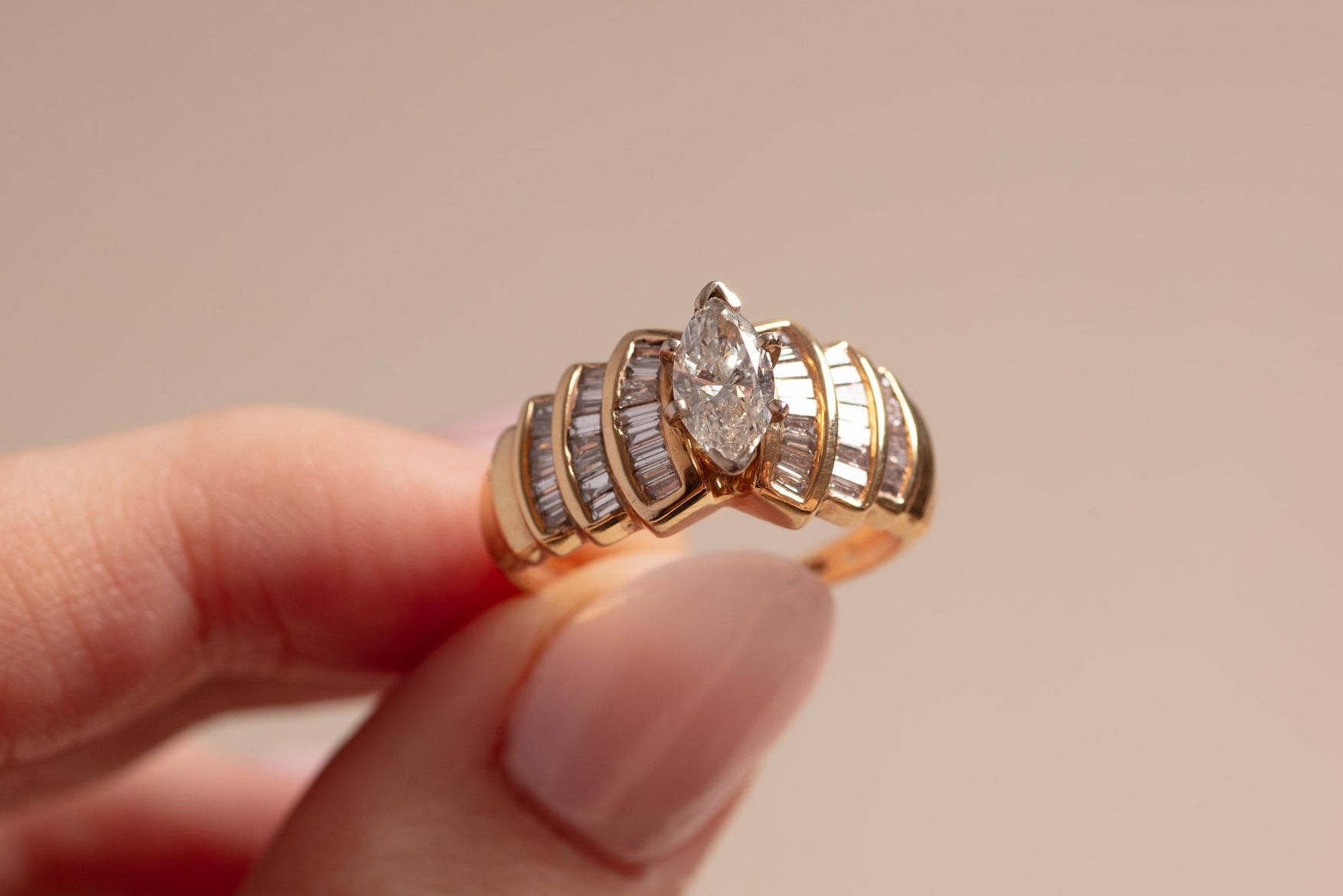 Gold ring with a marquise diamond held between fingers against a beige background